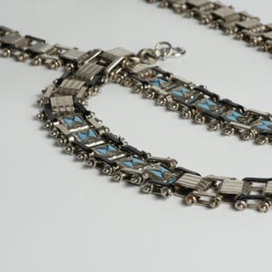 May include: A silver-toned metal chain necklace featuring a repeating geometric pattern. The design incorporates alternating rectangular silver links, smooth black enamel accents, and small triangular light blue enamel inlays throughout.