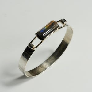 May include: A silver bracelet with a rectangular, iridescent stone at the center. The stone displays a spectrum of colors. The bracelet has a simple, modern design with a hinged clasp.