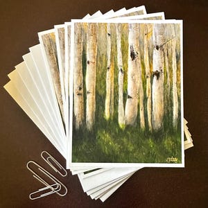 May include: A stack of square art prints featuring a forest scene with white birch trees and green grass. The prints have white borders and are accompanied by a stack of blank white cards and silver paper clips.