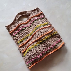 May include: A crocheted tote bag with a brown base and colorful stripes of pink, orange, and yellow. The bag has a woven handle.