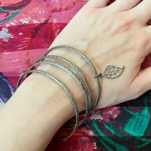 Könnte beinhalten: Ein silbernes Armband mit einem Blattanhänger. Das Armband besteht aus mehreren dünnen Bändern und wird an einem Handgelenk mit schwarzem Nagellack getragen.