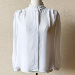 Puede incluir: Una blusa blanca de manga larga con cuello alto tipo mandarín, tapeta de botones oculta y hombros fruncidos. La blusa tiene mangas largas con puños abotonados y un dobladillo recto, presentada sobre un maniquí de color dorado.