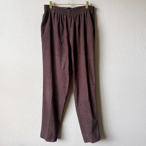 May include: A pair of brown pants with an elastic waistband hangs from a gold-colored hanger. The pants are made of a soft, textured fabric and have a straight leg design. The pants are a solid color and appear to be vintage.