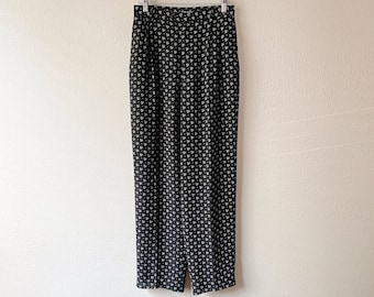 Puede incluir: Pantalón negro con estampado floral blanco colgado de una percha de madera contra una pared blanca.