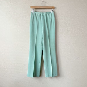 Pode incluir: Uma calça de perna larga verde claro com textura canelada. A calça está pendurada em um cabide de madeira contra uma parede branca.