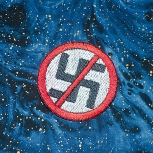 May include: Embroidered patch featuring a black swastika symbol crossed out with a red diagonal line, inside a red circle. The patch is on a blue fabric with a galaxy-like pattern and gold flecks.