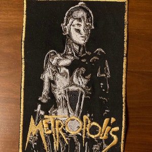 May include: Embroidered patch featuring a stylized image of a robot from the movie "Metropolis" in black, white, and gold. The word "Metropolis" is embroidered in gold script below the robot figure. The patch has a gold border.