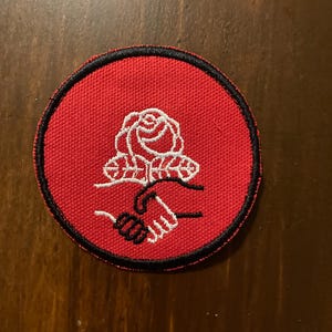 May include: A round, red patch with a black border. The patch features a white rose above two clasped hands, also in white. The patch is likely an emblem or logo, possibly for a club or organization.
