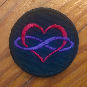 Pode incluir: Um remendo bordado preto com um coração vermelho e um símbolo de infinito roxo dentro do coração.