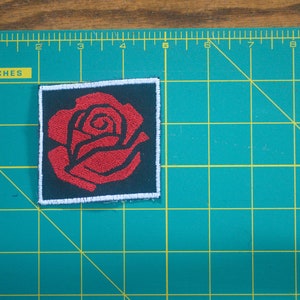 May include: A red embroidered rose patch with a white border and black background.