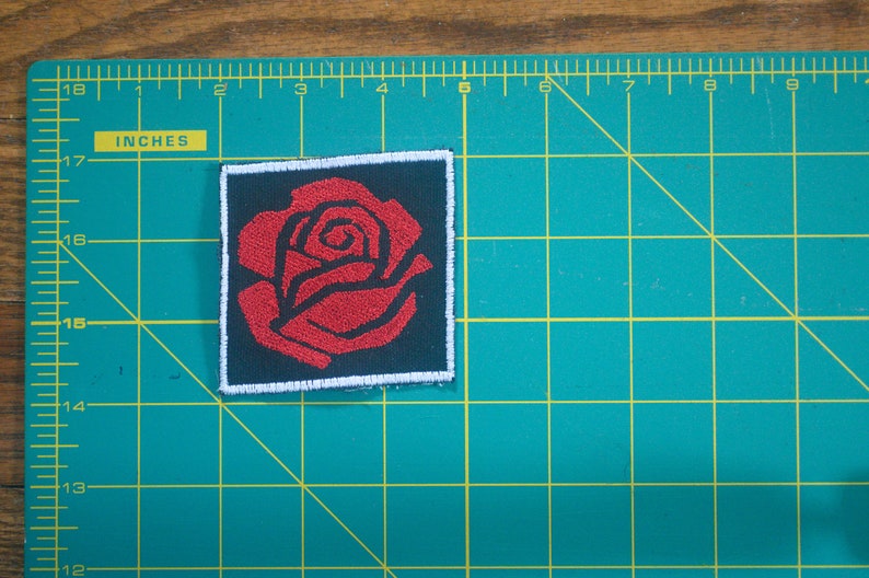 May include: A red embroidered rose patch with a white border and black background.