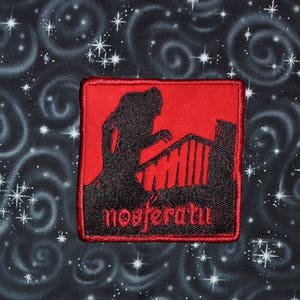 Peut inclure: Un écusson rouge brodé avec une silhouette noire d'une figure sur un balcon, avec le mot "nosferatu" en script rouge. L'écusson carré est posé sur un tissu noir avec des étoiles blanches et des motifs tourbillonnants.