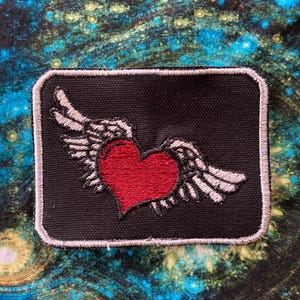 May include: A black fabric patch with a red heart and gray wings. The patch is surrounded by a gray border.