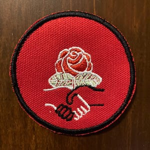 May include: A round, red patch with a black border. The patch features a rose with green leaves and two hands shaking. The rose is red and white, and the hands are black and white.