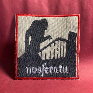 May include: A black and white embroidered patch with a silhouette of a figure climbing a staircase. The patch has a red border and the word "noefratu" is embroidered in white on the bottom.