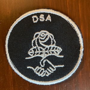 May include: Black and white embroidered patch with the letters "DSA" at the top. The patch features a rose and two hands shaking in the center, with a white border.