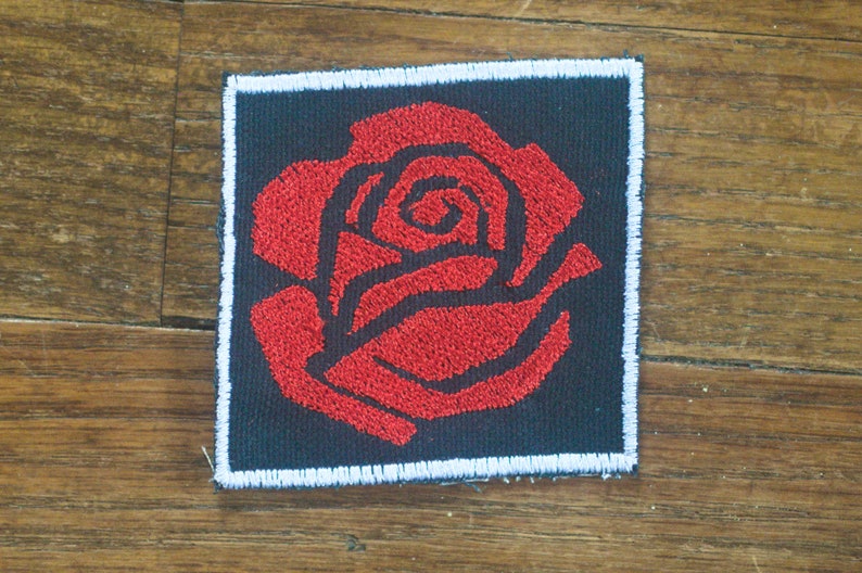 May include: A red embroidered rose patch on a black background with white stitching around the edges.