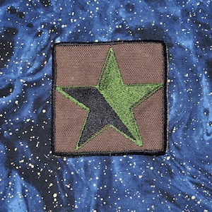 May include: A brown fabric patch with a black and green star. The star is a five-pointed star with the points of the star overlapping.