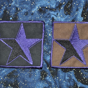 Op de afbeelding: Twee stoffen patches met een paarse ster op een zwarte en bruine achtergrond. De ster is omrand met paars en heeft een zwart midden.