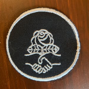 May include: Circular black patch with a white embroidered design. The design features a rose above two clasped hands, with a white border. The patch is likely for clothing or accessories.