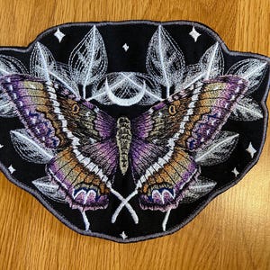 Może przedstawiać: Haftowana naszywka z motywem motyla w kolorach fioletu, brązu i złota na czarnym tle. Naszywka zawiera białe akcenty, półksiężyc i gwiazdy, idealna do kurtek lub toreb.