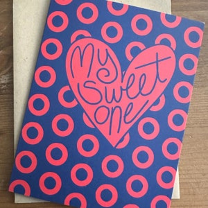 May include: A blue card with a pink heart shape and the text "My sweet one" inside the heart. The background is a pattern of pink donuts on a blue background.