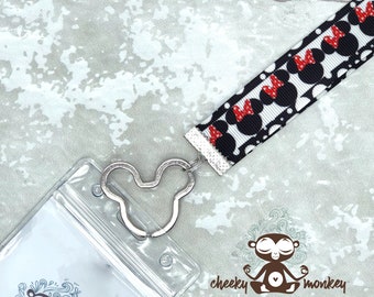 May include: A clear plastic badge holder with a Mickey Mouse shaped metal clip attached to a black and white ribbon with red polka dots and red Mickey Mouse heads. The ribbon is attached to a silver swivel clip. The badge holder has a clear plastic window for displaying a badge or ID card. The text "cheeky monkey supply company" is printed on the bottom of the image.