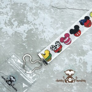 May include: White lanyard with a clear plastic badge holder. The lanyard features a repeating pattern of colorful cartoon Mickey Mouse heads. The badge holder has a silver Mickey Mouse shaped clip. The lanyard has the text "cheeky monkey supply company" printed on it.