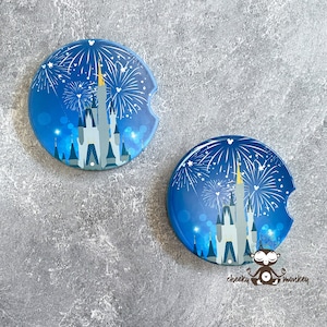 May include: Two round car coasters with a blue background featuring a castle and fireworks design. The castle is gray with a yellow spire. White fireworks burst across the top. The coasters have a small cutout for easy removal.