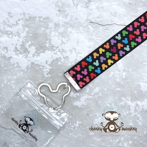 May include: A black lanyard with a rainbow Mickey Mouse pattern. The lanyard has a clear plastic badge holder with a Mickey Mouse shaped clip. The lanyard is attached to the badge holder. The lanyard has the text "cheeky monkey supply company" on it.