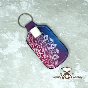 May include: A lip balm holder with a key ring. The holder is a gradient of purple and blue, with a white mandala design. A white lip balm is inserted in the holder. The logo of "Cheeky Monkey Supply Company" is in the bottom right corner.