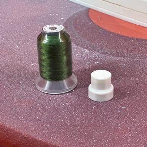 May include: A spool of dark green thread with a clear plastic base and a small white plastic cap. The thread is tightly wound, and the cap is cylindrical. The items are on a textured, reddish-purple surface.