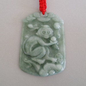 May include: A jade pendant featuring a carved monkey holding a peach, suspended on a red string necklace. The pendant is green and intricately carved with the monkey and peach.