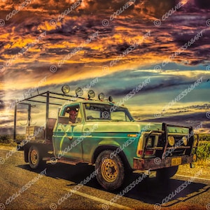 May include: A vintage blue pickup truck with a wooden bed drives down a paved road. The truck has a roll bar and a driver in the cab. The sky is a vibrant orange and purple sunset with clouds.