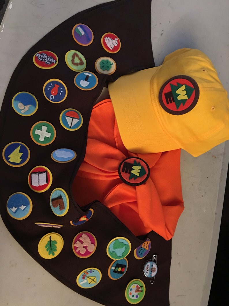 May include: A brown sash with a variety of colorful patches, including a first aid cross, a book, a leaf, and a globe. The sash is worn with a yellow baseball cap with a brown patch that says "W" and a tree.