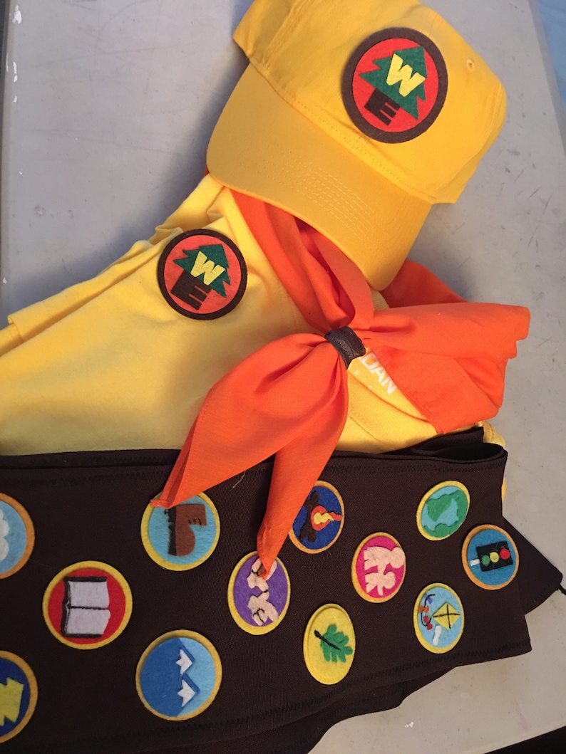 May include: A yellow scout uniform with a brown sash featuring various merit badges. The sash has a green, brown, and white tree emblem with the letter "W" in the center. The uniform also includes a yellow cap with a matching tree emblem and an orange neckerchief.