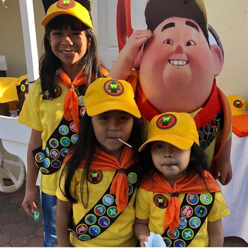 May include: Three children dressed in yellow scout uniforms with orange sashes and badges. They are wearing yellow hats with a green tree and a W on them. The child in the middle is holding a lollipop. A cartoon character of a scout leader is in the background.