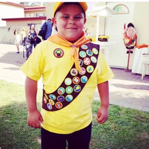 May include: A young person dressed as a Wilderness Explorer from the movie Up. They are wearing a yellow shirt with a brown sash and many badges. The sash has a  "W" shaped badge with a tree on it. The person is wearing a yellow hat and has a friendly expression.