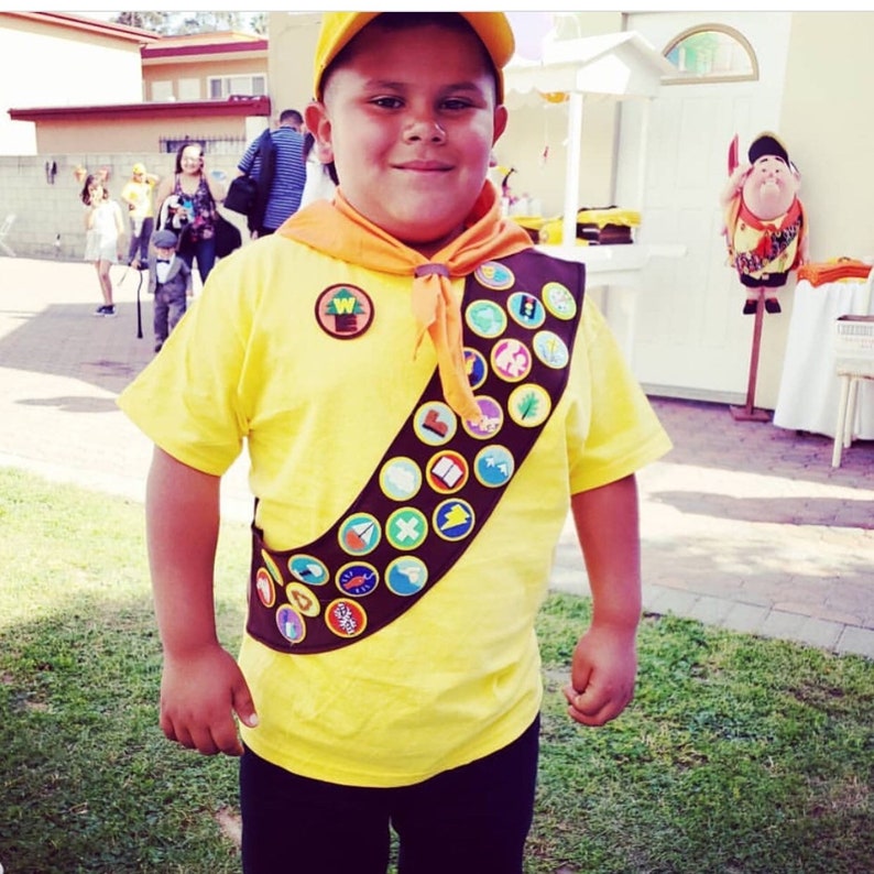 May include: A young person dressed as a Wilderness Explorer from the movie Up. They are wearing a yellow shirt with a brown sash and many badges. The sash has a  "W" shaped badge with a tree on it. The person is wearing a yellow hat and has a friendly expression.