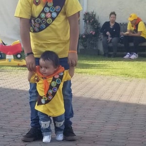May include: A father and son dressed as characters from the movie "Up" wearing yellow shirts with badges and orange sashes. The boy is holding his father's hand.
