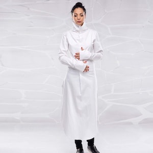 May include: A white long-sleeved tunic with a high collar and long sleeves. The tunic has a relaxed fit and falls to the ankles. The model is wearing black pants and black shoes.