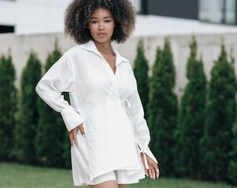 May include: A woman wearing a white linen shirt dress with a tie belt and white sandals. She is standing by a swimming pool.