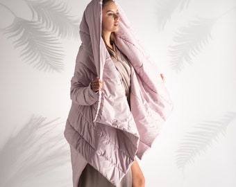 Könnte beinhalten: Ein helllila, gesteppter Kapuzenmantel mit langem, fließendem Design. Der Mantel wird über einem langen, taupefarbenen Kleid getragen. Das Model trägt High Heels. Der Hintergrund ist weiß mit einem Palmenblattschatten.