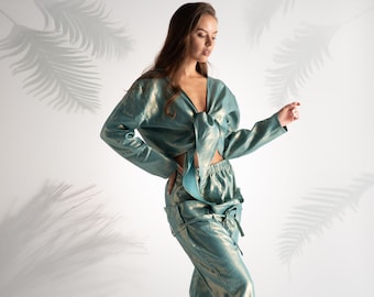 Puede incluir: Un conjunto de dos piezas de color verde azulado con un brillo metálico. El conjunto incluye un top de manga larga con lazo y pantalones a juego de cintura alta. La modelo lleva sandalias blancas de tacón. Fondo blanco con sombras de hojas de palmera.