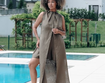 May include: A woman wearing a long, olive green linen dress with a high neckline and a slit in the front. She is standing near a swimming pool and has her hair pulled back.