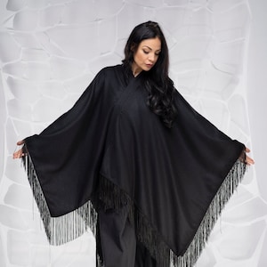 Puede incluir: Un poncho negro con flecos. El poncho se lleva sobre pantalones negros y la modelo lleva tacones negros.