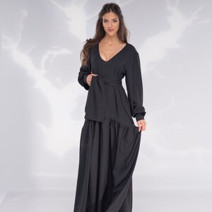 May include: A woman wearing a long black dress with a V-neckline and a tie at the waist. The dress has a flowing skirt and long sleeves.