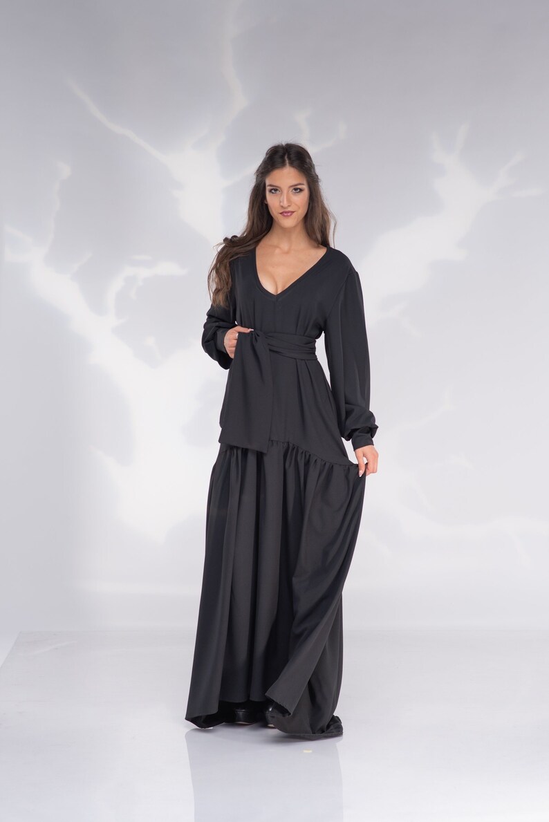May include: A woman wearing a long black dress with a V-neckline and a tie at the waist. The dress has a flowing skirt and long sleeves.