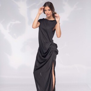 May include: A floor-length black dress with a draped design and a high side slit. The dress features a boat neckline and short sleeves. The model is wearing black high heels.