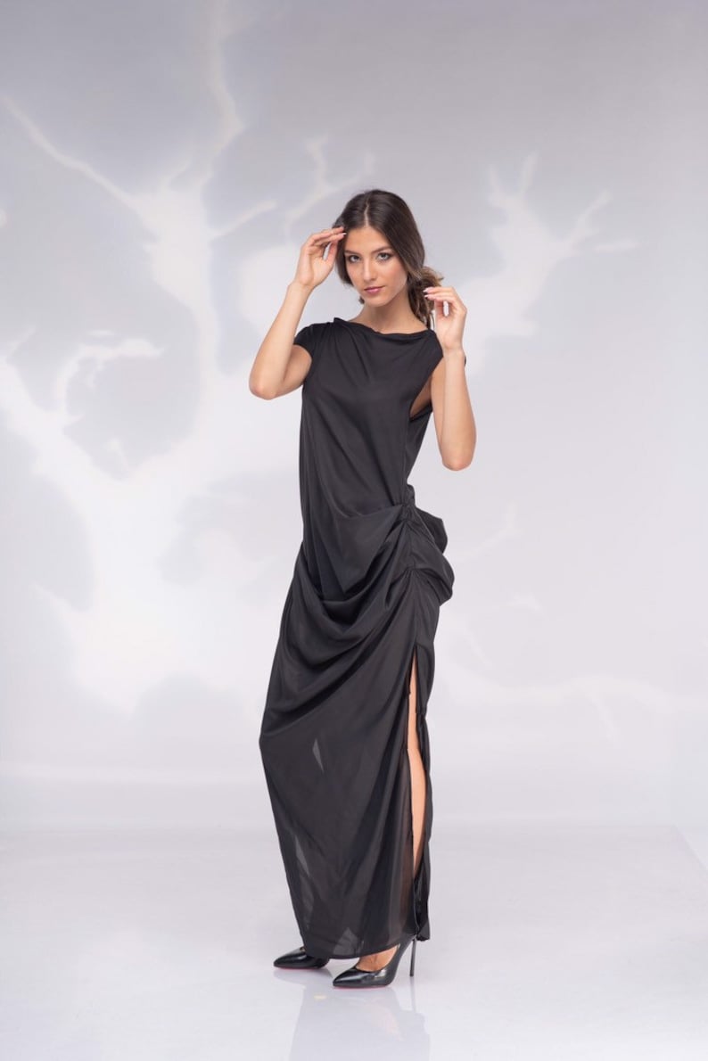 May include: A floor-length black dress with a draped design and a high side slit. The dress features a boat neckline and short sleeves. The model is wearing black high heels.
